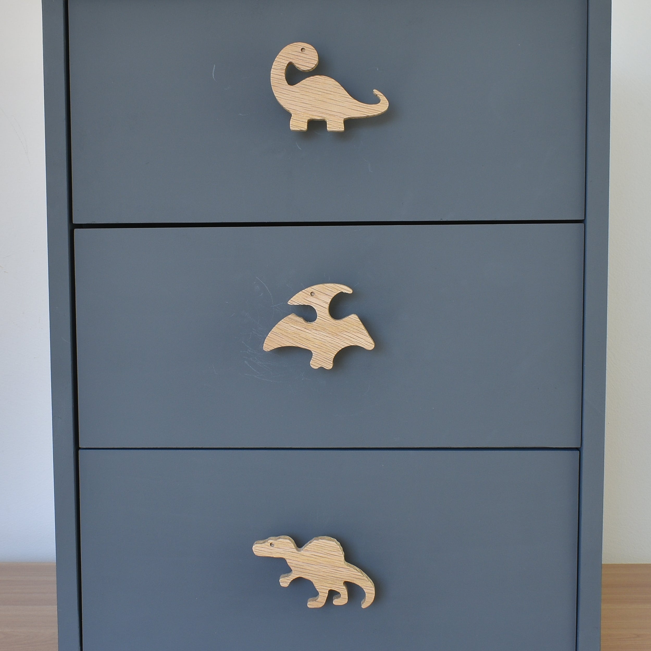 Oak dinosaur drawer knobs fitted to a blue chest of drawers showing brachiosaurus, pterodactyl and spinosaurus designs