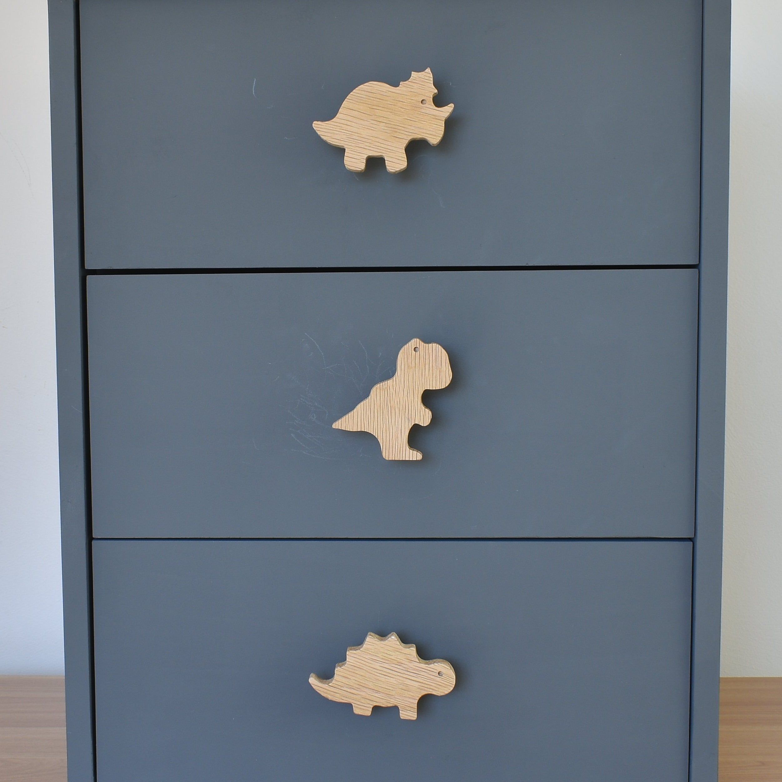 Oak dinosaur drawer knobs fitted to a blue chest of drawers showing triceratops, T rex and stegosaurus designs